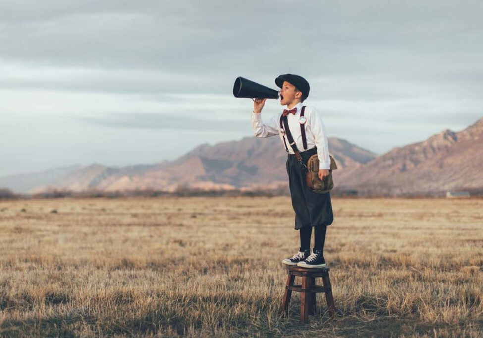 A news boy dressed in vintage knickers and newsboy hat stands yelling through a megaphone in the middle of a field in Utah, USA. He is trying to sell you what your business needs.